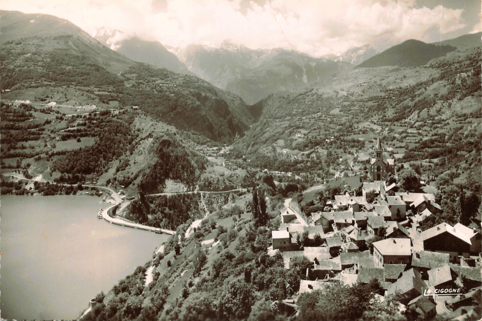 Une vue du village de Mizoën depuis les hauteurs, avec le lac en contrebas et une végétation naissante, sans date précis