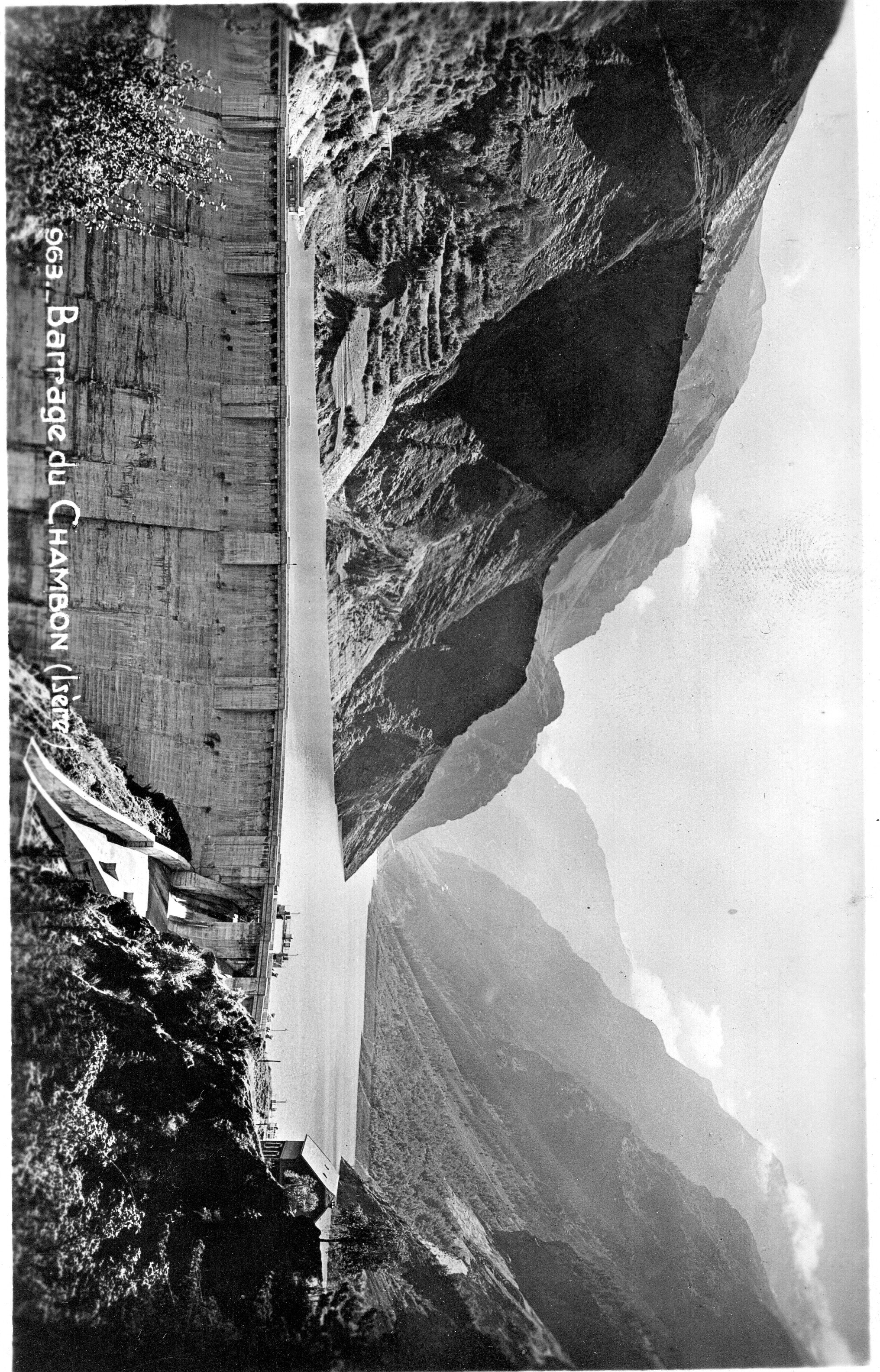 Une vue du barrage depuis l'aval, avec les massifs d'Emparis et des Ecrins en arrière-plan, offrant un contexte géograph