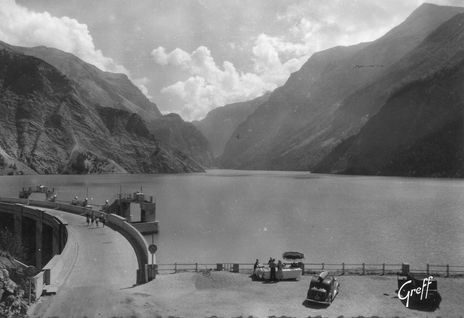 Des voitures garées devant le lac du Chambon, avec des piétons sur le barrage et un stand de vente de boissons. Cette sc